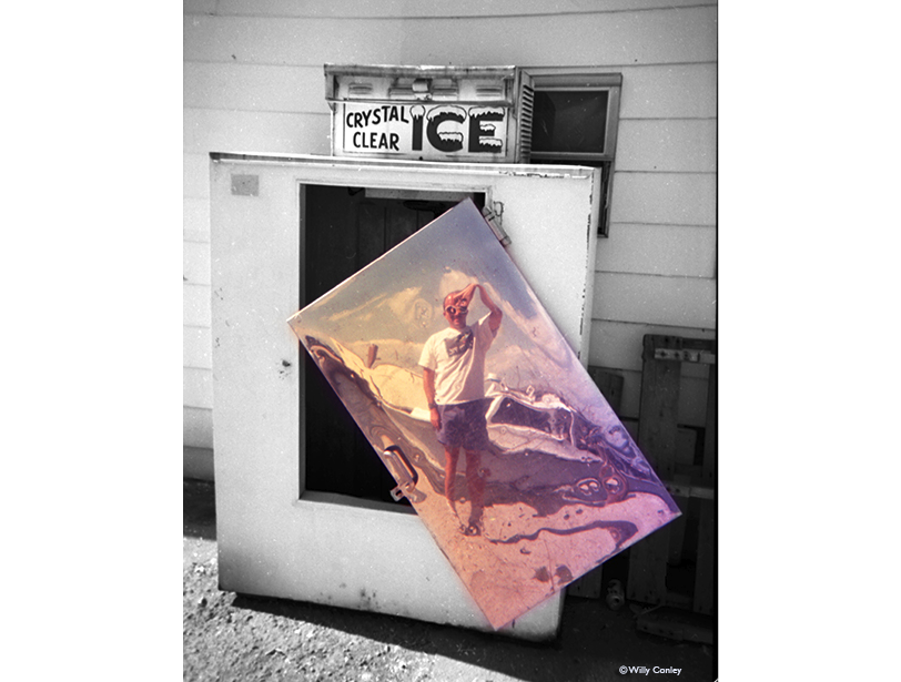 black-and-white and color vertical photo of a dilapidated ice machine with its shiny door hanging sideways held up by a screw. On top of the machine is the condenser with the machine’s advertising label: “Crystal Clear ICE”. On the door is a warped reflection, in color, of the author, in a white t-shirt and blue shorts holding a camera over his face. Behind him is the image of a fishing boat. The tone of everything surrounding the hanging door is black and white.