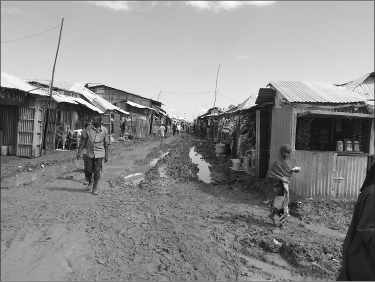 A muddy street in Kakuma Refugee Camp, lined with corrugated iron shacks. Some have bright plastic buckets and chairs stacked outside them, indicating that they are shops; there is also a basic kiosk with a serving hatch. There are people in the distance; in the foreground, a black man in muddy clothes and wellington boots is walking towards the camera followed by other men, and a young black girl with bare feet and a purple headscarf is crossing in front of the kiosk, carrying a small paper bag. 