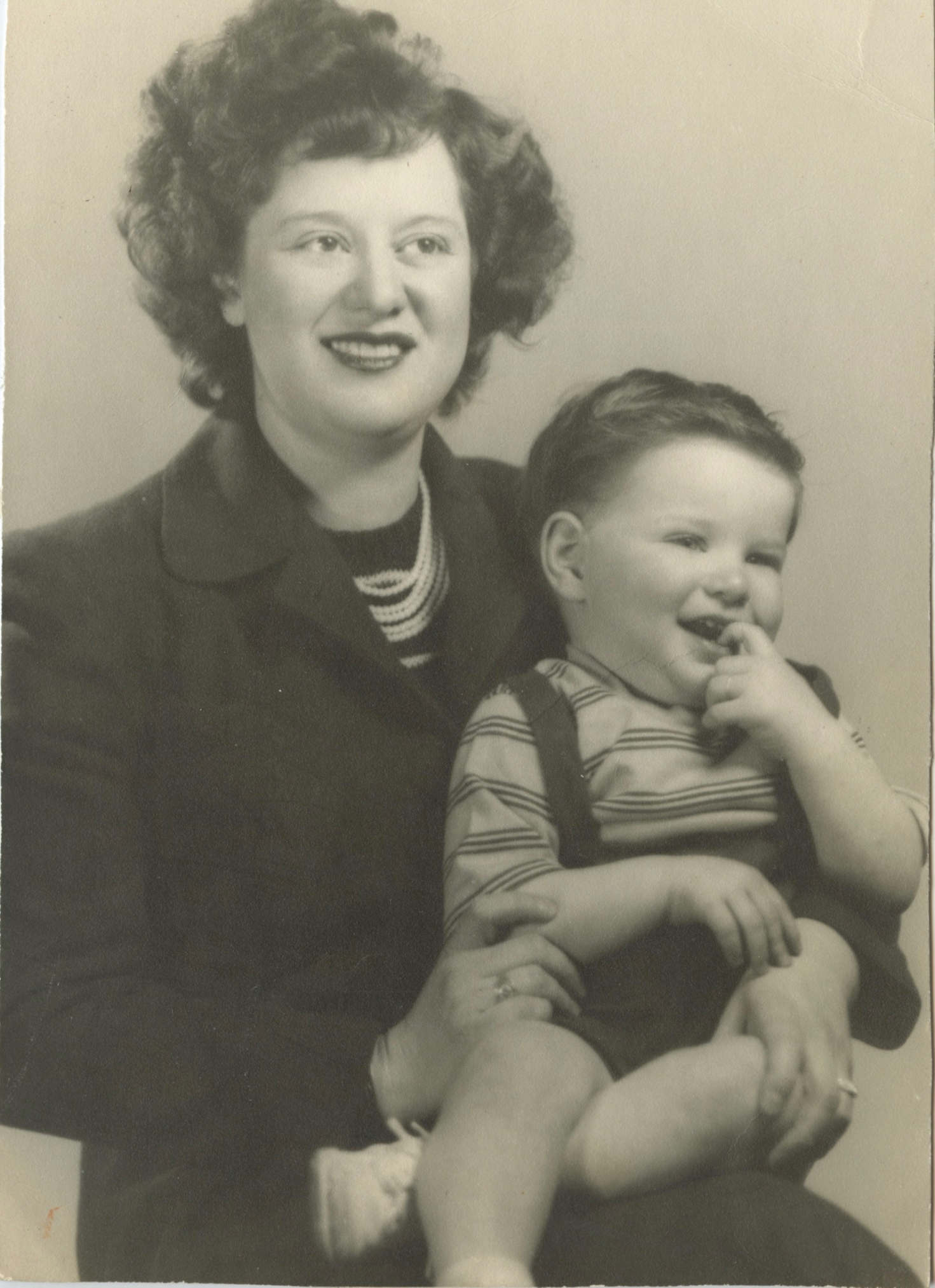 A young white woman with wavy dark hair holding a smiling little white boy who has his index finger in his mouth. They are both looking off to the right.
