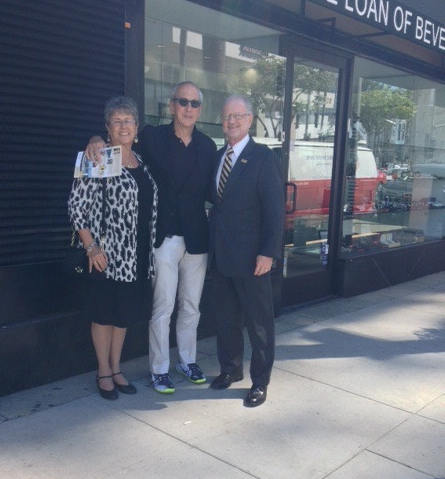 Three older white adults stand on the sidewalk in front of a bank. The woman is wearing a black-and-white jacket with a cheetah spots pattern over a black dress and black flat shoes. The man next to her is tall and slim and has his arm around her. He's wearing sunglasses, a dark jacket, white pants and sneakers. The man next to him is shorter and is wearing a navy suit with a striped tie.