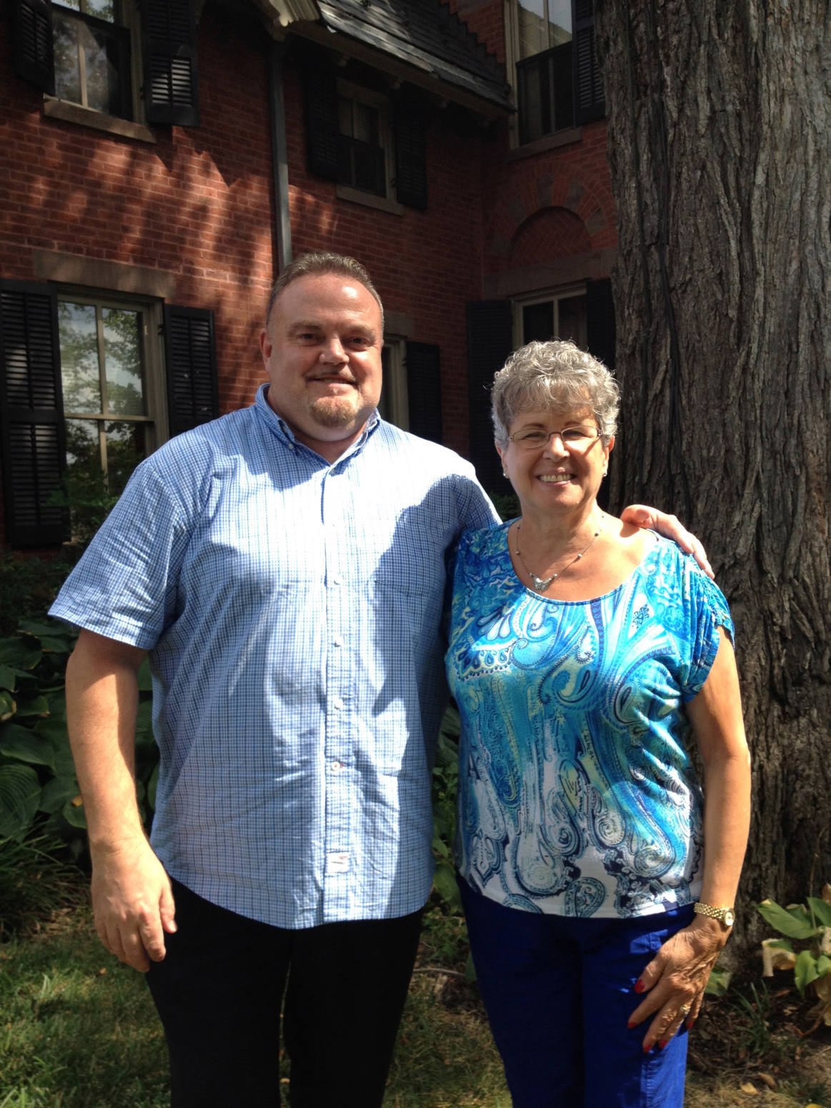 A middle-aged white man with a goatee and short hair in a blue checked button-down shirt stands next to an older white woman with gray wavy hair and glasses. She's wearing a blue top with a swirled pattern. They are outside in front of a brick house.