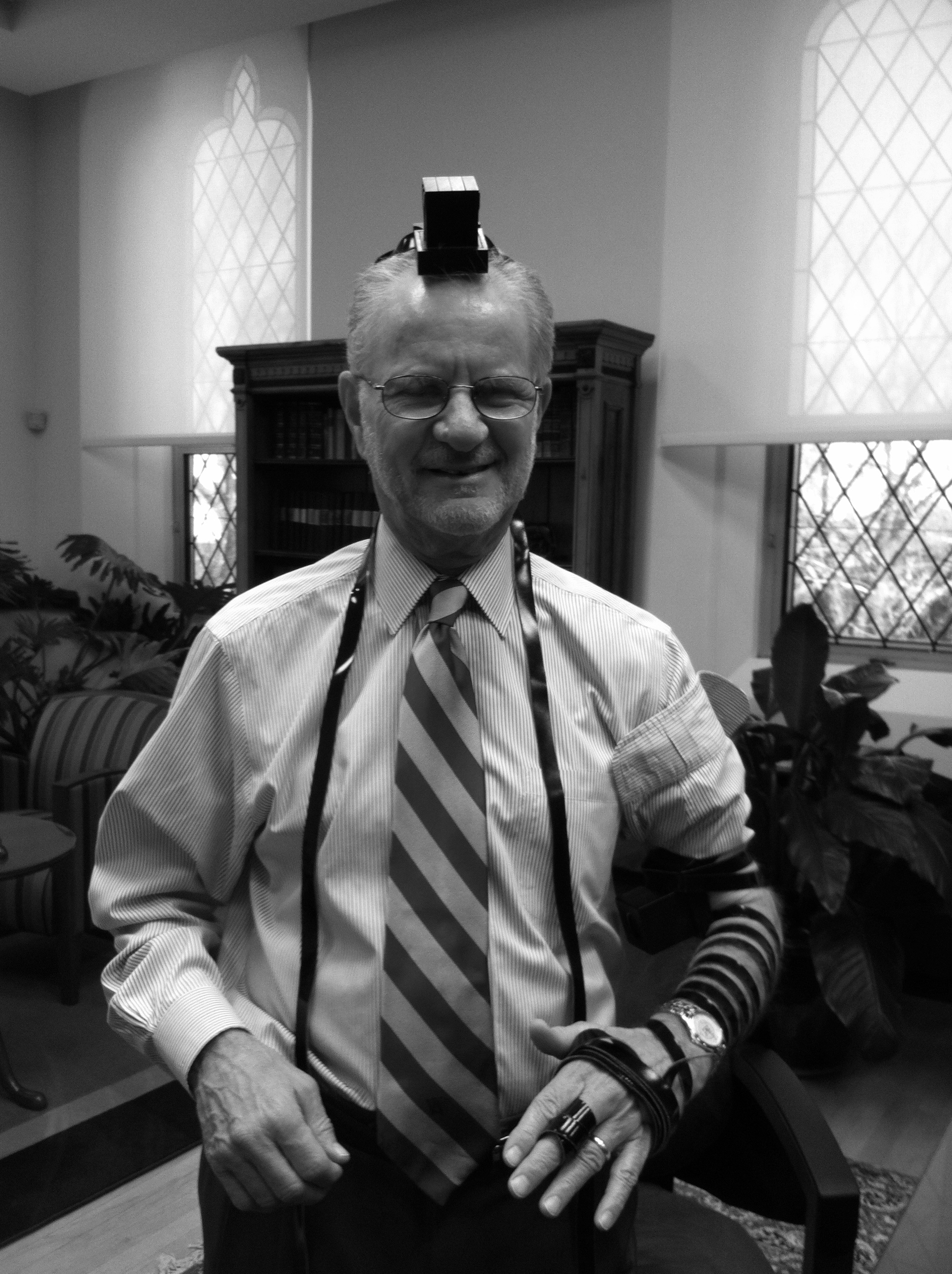 An older white man with glasses smiles in to the camera while wearing Tefillin in an office.