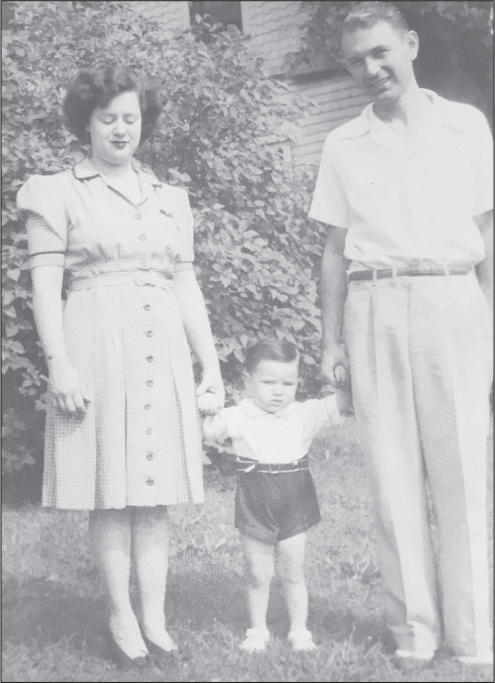A black-and-white photo of a young white woman with dark wavy hair in a light-colored dress with buttons down the front. Her eyes are closed. She's holding the hand of an unsmiling little white boy with short hair in shorts. A young smiling white man holds the other hand of the toddler. He's wearing a white collared short-sleeve shirt and long pants with a black belt. Behind them is a large bush and a house.