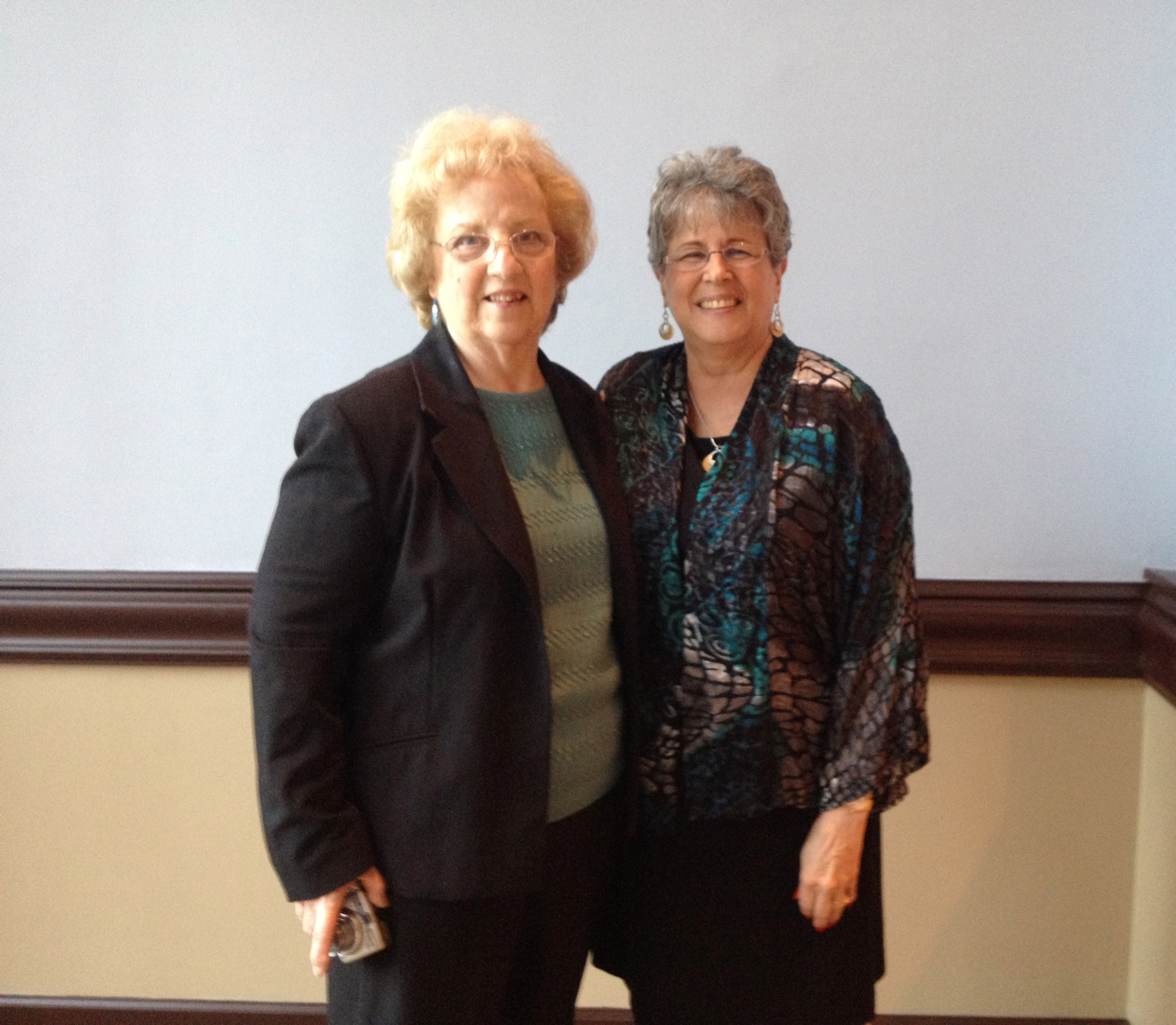Two older white women stand together. The one on the left has curly blond hair and glasses. she's wearing a black jacket over a green top. Next to her is a slightly shorter woman with gray wavy hair and glasses.