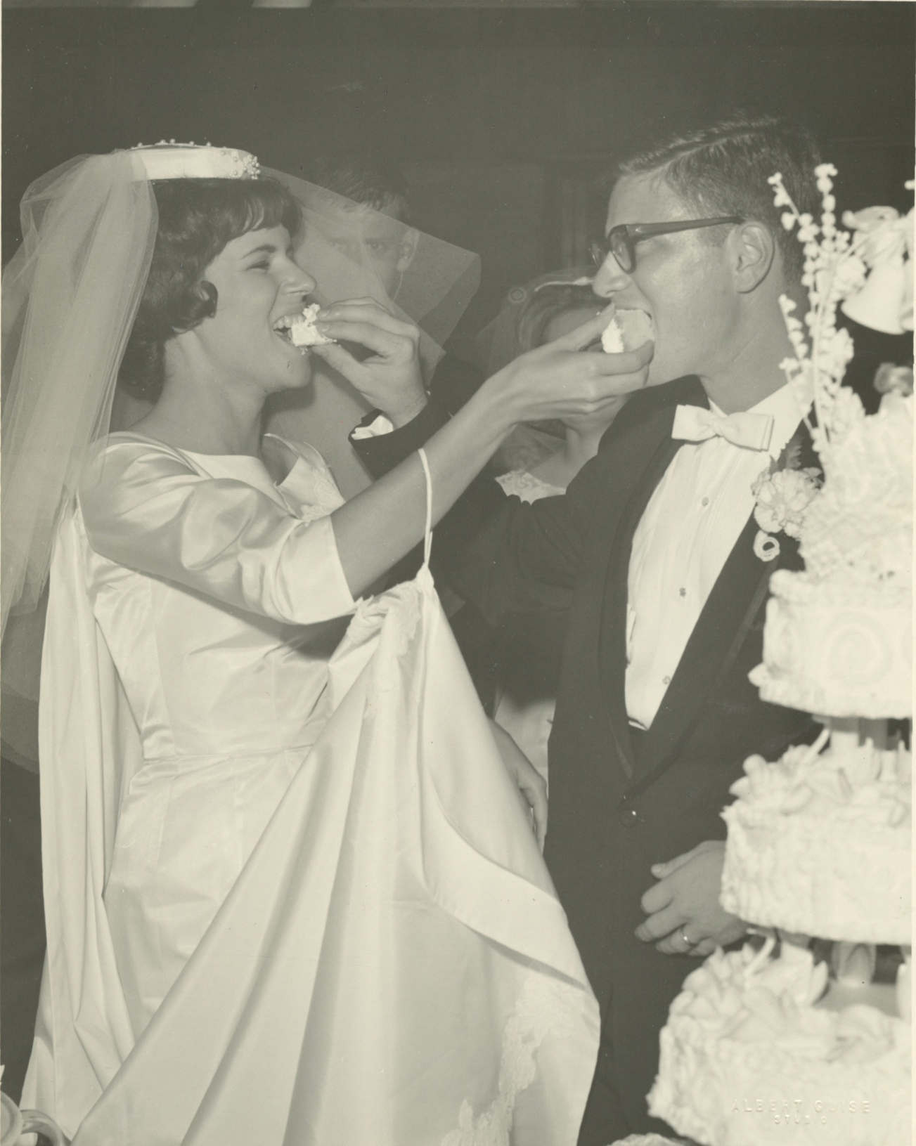 A young white woman in a white wedding dress and veil feeds a slice of cake to a young white man in glasses and a tuxedo. He is also feeding her a slice at the same time. The large, multitiered wedding cake is in the foreground.