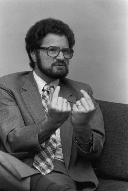 Black and white photo of a young white man wearing a suit and a plaid tie. He has dark rimmed glasses, dark hair and a dark beard and mustache. Both of his arms are up and both of his index fingers are pointing back at himself.