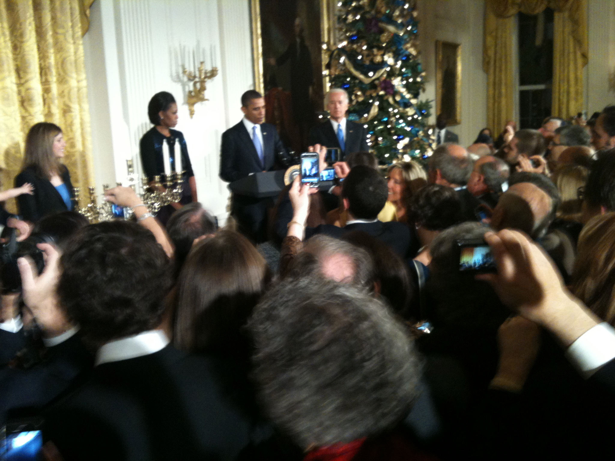 A tall Black woman (Michelle Obama) in a dark top stands underneath a gold candelabra mounted on a white wall. Next to her, and slightly in front of her, a tall Black man (President Obama) in a dark colored suit with a light blue tie stands at a podium and is speaking into microphones. On the other side of him stands a white man in a dark colored suit with a slightly bluer tie (Vice President Biden). Behind him is a large portrait of George Washington  as well as a very large Christmas tree decorated in blue and gold. There is a crowd in front of the podium of mostly white people.