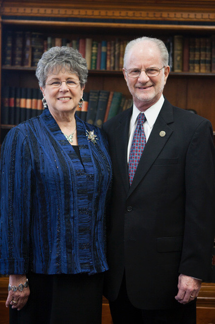 An older couple, a white man and woman, stand in front of a bookshelf. The woman is wearing a blue-and-black top, silver hoop earrings, a silver necklace, and a large silver brooch. She has short salt and pepper hair. The man is wearing a black suit with a light blue and red tie. He has a gold pin on his lapel. Both are smiling for the camera.