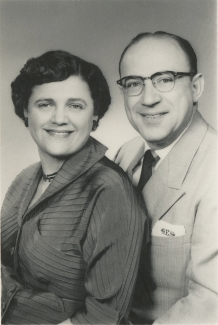 Black and white photo of a middle aged man and woman. The woman has short dark hair, parted on the side. She is wearing a dark striped dress with buttons down the front. The man is balding and is wearing a light colored suit jacket. His glasses have dark half frames. In his suit chest pocket he has a handkerchief mongrammed with an E. Both are smiling at the camera.