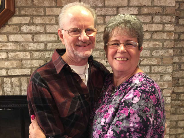 An older man and woman stand in front of light colored brick fireplace. The man is wearing a reddish brown and black plaid buttoned up shirt with a white t-shirt underneath. The woman has short salt and pepper hair and is wearing a top with pink and light pink flowers on a black background. She has silver hoop earrings and a silver necklace. They are holding each other but are turned, smiling, at the camera.