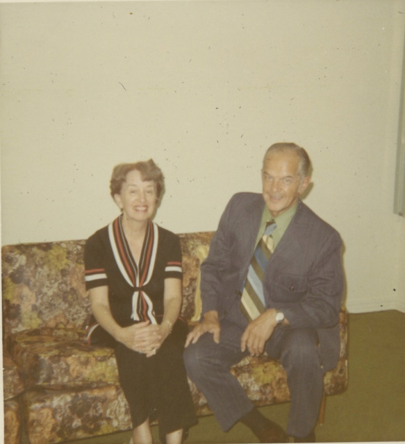 A middle aged white man and woman sit on a brown and yellow floral patterned couch. The woman has her legs crossed and her hands are clasped around her knee. She has on a black dress with red and white accents around the neckline and the cuffs of its short sleeves. She has short brown wavy hair. The man is in a gray suit with a green shirt and a wide, striped tie. He has receding white hair and a tan.