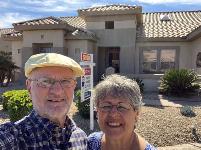 An older white man and woman, smiling, standing in front of a one-story beige and tan house with tan terracotta roof tiles and a sold sign out front. The man is smiling, wearing a yellow cap and a dark blue-and-white checkered shirt. The woman is also smiling and is wearing a blue and white top.