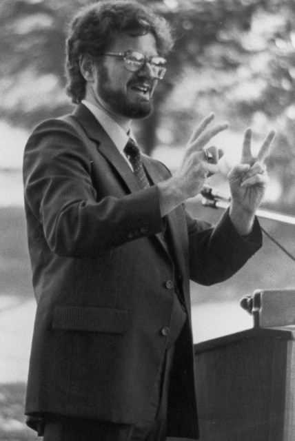 Black and white photo of a young white man standing at a podium. He has glasses, dark hair, and a dark beard and mustache. He is wearing a suit. His hands are raised and his index and middle fingers on both hands are extended in a V shape.