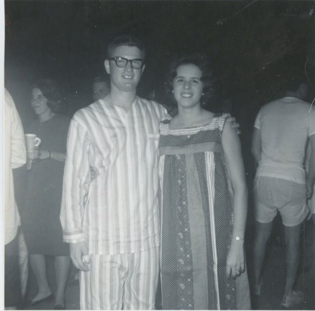 Black-and-white photo of a young white man and woman. The man is dressed in vertically striped pajamas. He has large black glasses and a short hair cut. His arm is around a young woman with wavy chin-length hair, wearing a sleeveless vertically striped housecoat. 