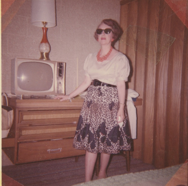 A young woman stands in front of a dresser in a room. On top of the dresser is an old-fashioned television with a lamp on top of it. The woman is dressed in a white blouse with puffy short sleeves, a black belt and a leopard spotted black and white skirt. She has a necklace made up of big orange beads with matching earrings. Her wavy reddish hair is chin length. She's wearing large dark sunglasses and red lipstick.